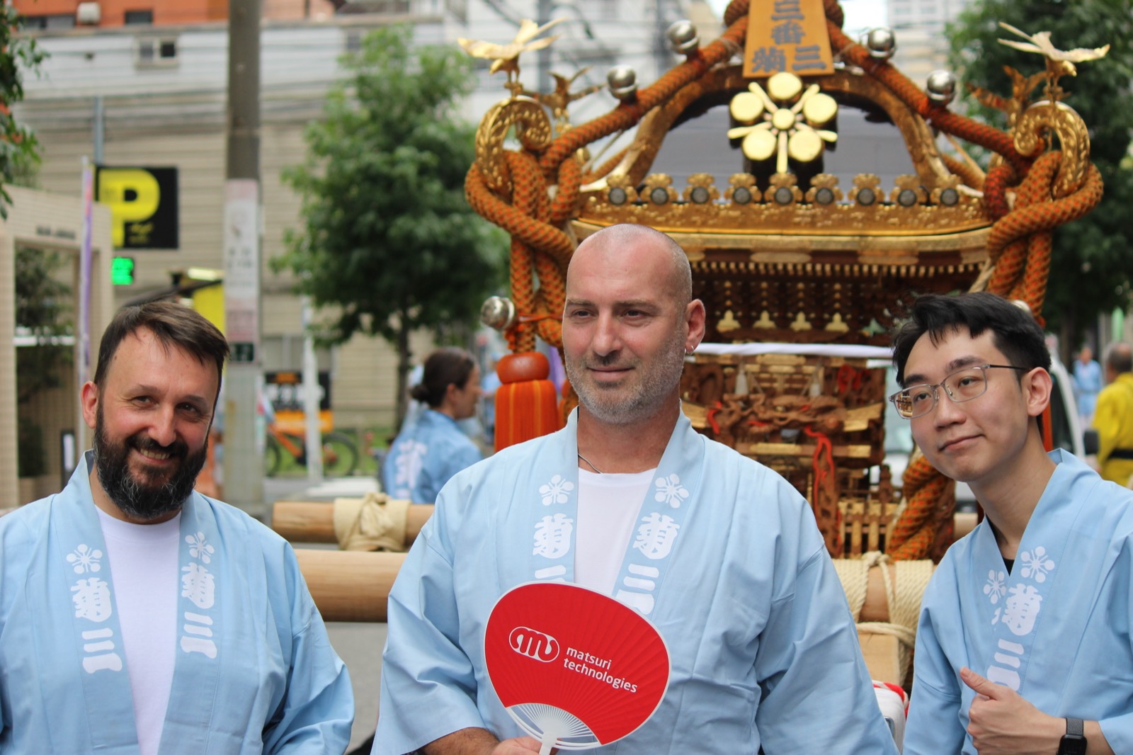 Three participants in happi coats in front of the mikoshi